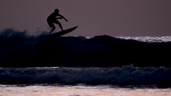 Silhouetted surfer riding a wave during golden hour with dramatic sky and ocean backdrop.