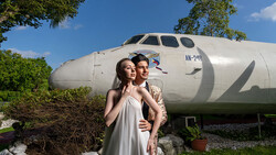 Couple posing in front of a large white aircraft fuselage in a garden setting.