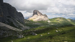 Alpine valley with steep rocky peaks and lush green meadows under cloudy sky.