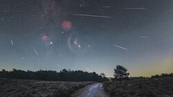 Meteor shower streaking across a twilight sky above a rural landscape with a stream and silhouetted trees.