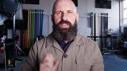 Man with full beard speaking to camera in a studio with photography equipment visible in the background.
