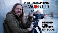 Photographer with long hair holding a medium format camera on tripod, smiling at camera in urban setting.