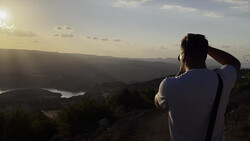 Photographer silhouetted against a vast landscape with mountains and a winding river at golden hour.