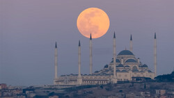 Full moon rising behind a mosque with four minarets and a central dome at dusk.