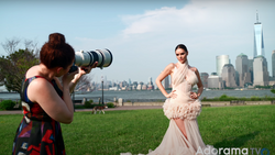 Photographer with telephoto lens capturing portrait of woman in pink gown with NYC skyline backdrop.