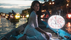 Woman in white clothing holding a decorative lantern in a boat at dusk among illuminated buildings.