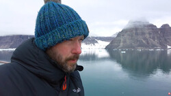 Man in blue knit beanie gazing toward snow-capped mountains across a glacial lake.