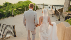 Bride and groom holding hands walking away on an outdoor stone terrace toward a railed overlook.