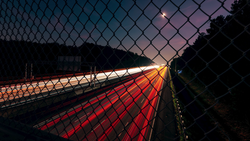 Highway at dusk viewed through chain-link fence, with red taillights creating light trails along the road.