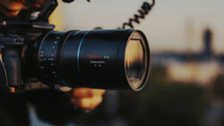 Close-up of a 150mm T2.9 anamorphic lens mounted on a camera, with a blurred cityscape in the background during golden hour.