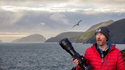 Wildlife photographer holding a telephoto lens while observing a seabird in flight over coastal cliffs.