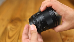 Hand holding a black camera lens with focus ring and depth-of-field markings visible.