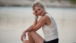 Woman in white knit tank top sitting on beach sand, smiling at camera.