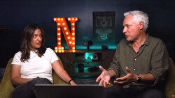 Two people in conversation during an interview or podcast recording set with neon signage in the background.