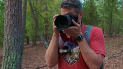 Photographer holding a telephoto lens camera in a forest setting, wearing a red shirt and backpack.