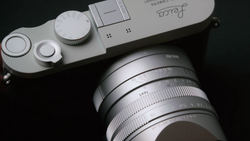 Silver mirrorless camera body with attached lens and visible control dials, photographed against a black background.