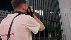 Photographer shooting upward at illuminated modern skyscraper at night with camera raised to eye level.