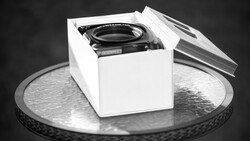 Camera lens in white product box on metallic circular surface against dark background.