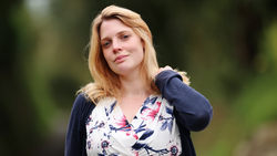Woman with blonde hair wearing a floral print blouse and navy cardigan, photographed outdoors with a soft, blurred green background.