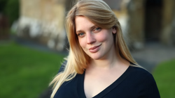Portrait of a young woman with long blonde hair wearing a black v-neck shirt, photographed outdoors with soft natural light.