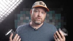 Man in baseball cap holding two cameras while speaking to camera in studio setting.
