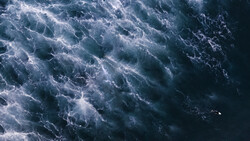 Aerial view of turbulent ocean water with white foam and wave patterns in dark blue-gray tones.