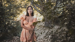 Woman in orange gingham dress posing among green foliage and tree trunks.