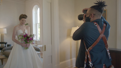 Photographer with camera on shoulder captures bride in white gown holding purple bouquet in elegant hallway.