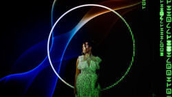 Person in green digital matrix-patterned dress standing within illuminated circular and curved neon light shapes against dark background.