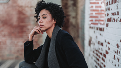 Woman with curly dark hair in thoughtful pose against weathered brick wall.