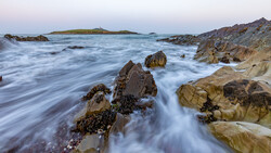 Rocky coastline with flowing water and distant island at twilight.