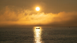 Fishing boat silhouetted against golden sunlight reflecting on calm ocean water at sunset.