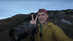 Photographer holding a Nikon camera with telephoto lens on a rocky hillside, making a peace sign at the camera.