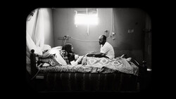 Black and white photograph of two people on a bed in a sparsely furnished room with bright overhead lighting.