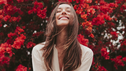 Woman with long brown hair smiling upward among vibrant red flowers in natural sunlight.