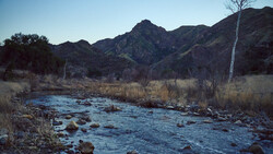 Mountain stream with rocks flowing through a valley with sparse winter vegetation and distant peaks.
