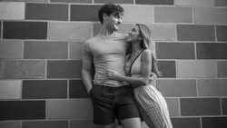 Couple posing against a brick wall, looking at each other with affection in black and white photography.