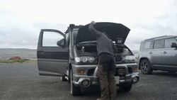 Photographer inspecting engine of parked vehicle during roadside shoot with overcast sky and mountains in background.