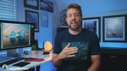 Man in dark t-shirt gesturing while speaking in a home office with blue ambient lighting and framed landscape photographs.