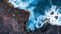 Aerial view of rocky coastline with waves crashing against dark stone cliffs.