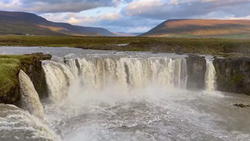 Powerful waterfall cascading over dark basalt cliffs into a turbulent river below, with mountains and moorland stretching across the background.