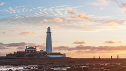 Lighthouse with keeper's cottage silhouetted against golden sunset sky with dramatic clouds.