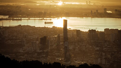 Golden-hour cityscape with dense urban skyline silhouetted against a bright waterway and hazy sky.