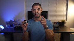 Man in blue t-shirt holding a camera in a home studio setting with blue and warm lighting.
