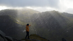 Hiker in yellow jacket standing on rocky ridge overlooking misty mountain valley.