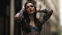 Tattooed woman with dark hair gazes upward in an urban alleyway, hands raised to her head.