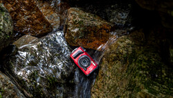 Red waterproof camera resting on moss-covered rocks beside flowing stream water.