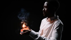 Person holding a lit candle against a dark background, with smoke rising from the flame.