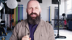 Bearded man in tan jacket gestures while speaking in a photography studio.