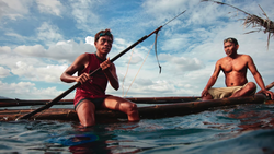 Two shirtless men in shallow water with wooden poles and a traditional boat.